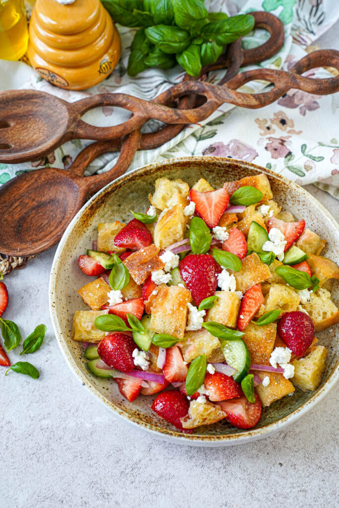 Close up of Strawberry Panzanella in a bowl next to basil leaves, wooden spoon, and wooden fork.