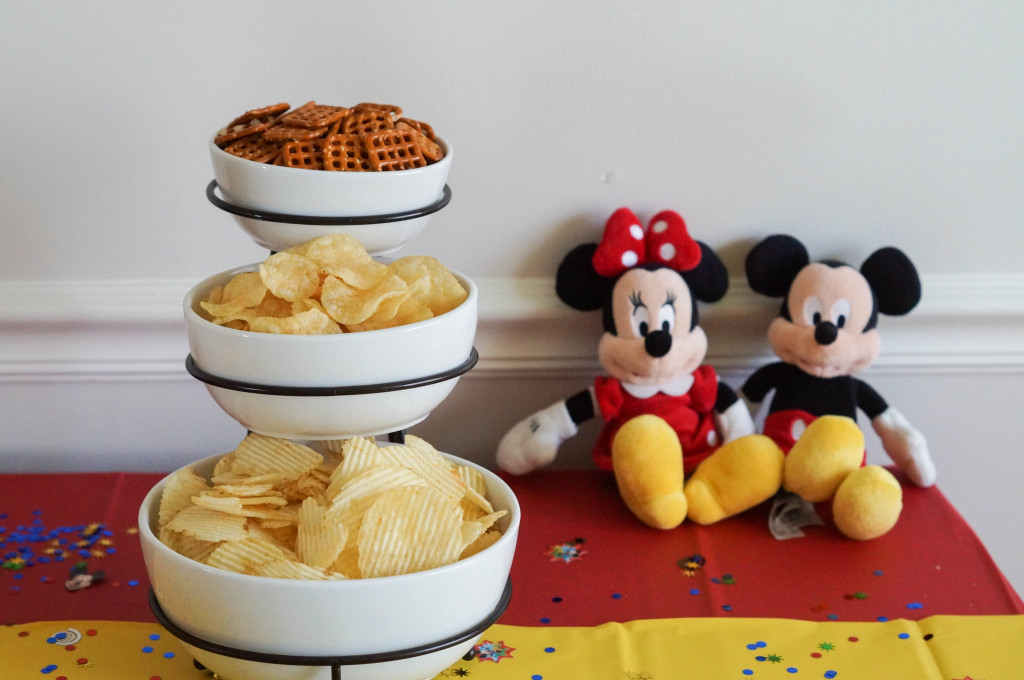 Chips and pretzels in white bowls next to stuffed Mickey and Minnie Mouse.