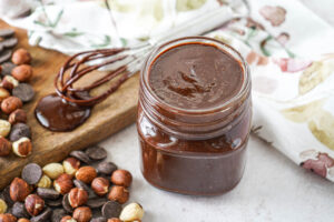 Crema Gianduia (Italian Chocolate Hazelnut Spread) in a jar next to a whisk, hazelnuts, and chocolate chips.