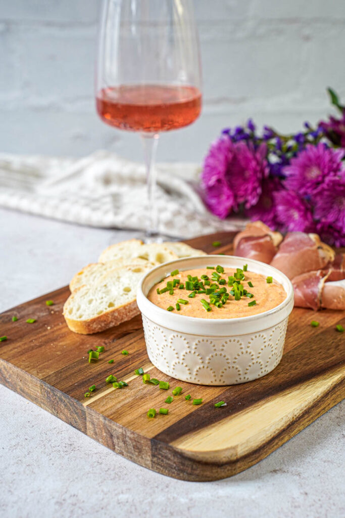 Side view of Liptauer (Austrian Cheese Spread) in a ramekin with flowers and a glass of wine in the background.