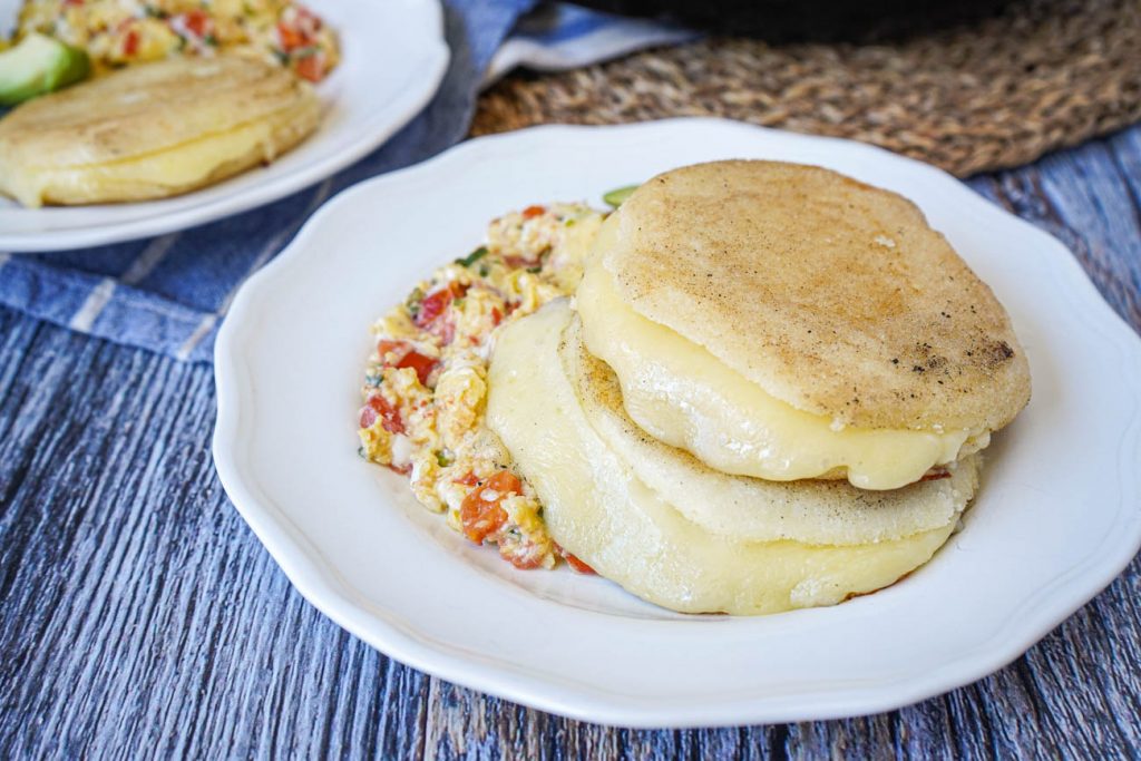 Two Arepas Rellenas de Queso (Cheese Stuffed Corn Cakes) on a white plate with scrambled eggs.