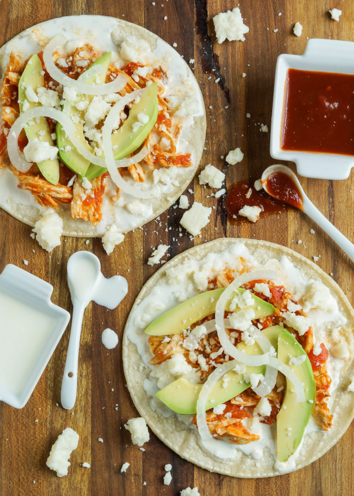 Aerial view of Chicken Tinga on two tortillas with scatter crumbled cheese, crema, chipotle sauce, avocado, and onion.