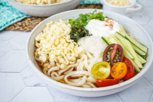 Hiyashi Tanuki Udon in a bowl with udon noodles, tempura flakes, tomatoes, cucumber, and egg.