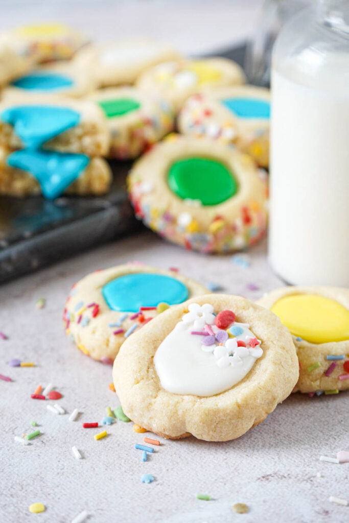 Close up of Iced Thumbprint Cookies with white, yellow, and blue icing in the center with more thumbprint cookies in the background.