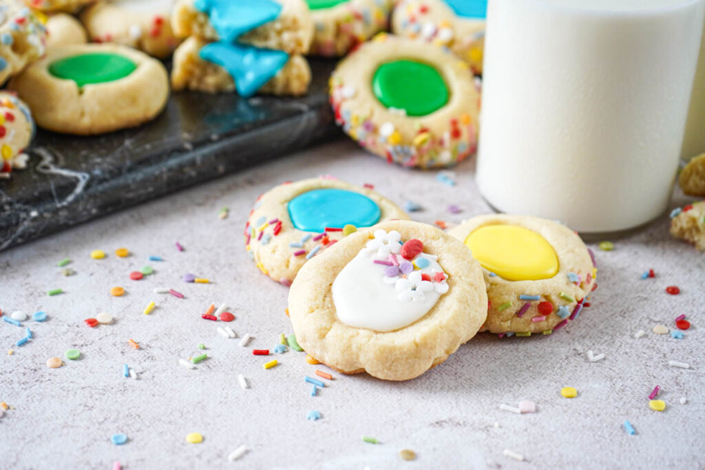 Iced Thumbprint Cookies with white, yellow, blue, and green icing in the center next to a glass of milk.