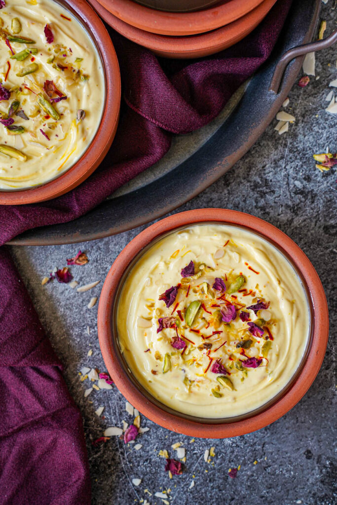 Aerial view of Kesar Elaichi Shrikhand (Indian Sweetened Saffron Cardamom Yogurt) in two clay bowls with pistachios, almonds, rose buds, and saffron.