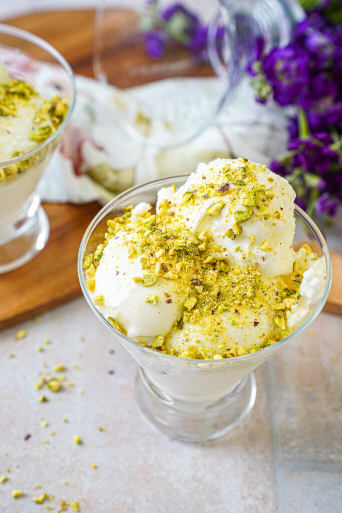 Mastic Ice Cream in a glass bowl covered in pistachios with another bowl and purple flowers in the background.