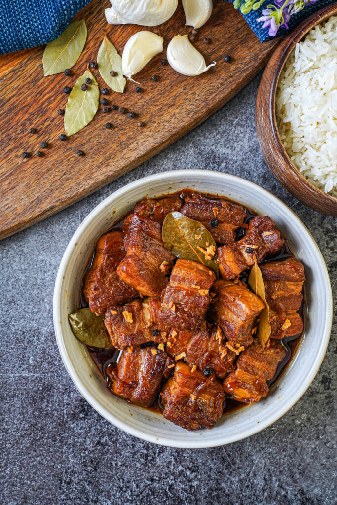 Aerial view of Adobong Baboy (Filipino Pork Adobo) in a bowl next to rice, bay leaves, peppercorns, and garlic cloves.