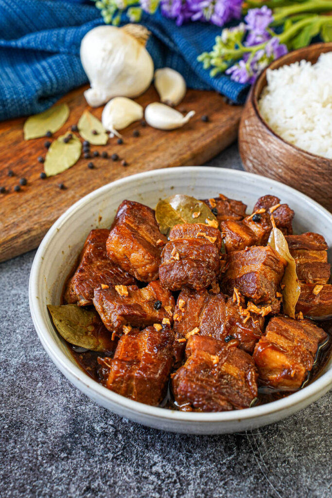 Close up of Adobong Baboy (Filipino Pork Adobo) in a bowl with rice, bay leaves, peppercorns, and garlic cloves in the background.