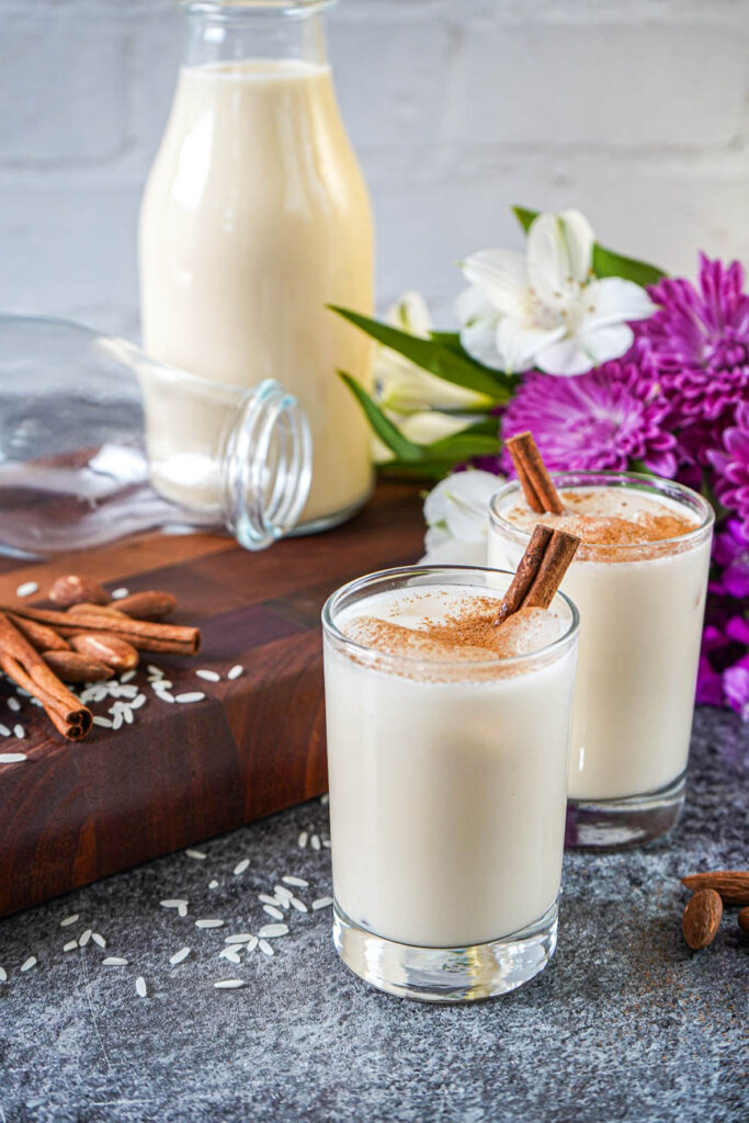 Side view of Horchata in two glasses with a pitcher and flowers in the background.
