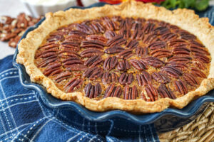 Pecan Pie in a blue pie dish with flowers and pecans in the background.