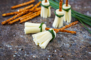 Two Pretzel Broomsticks on their side and three standing up in the background next to pretzel sticks and a bundle of chives.