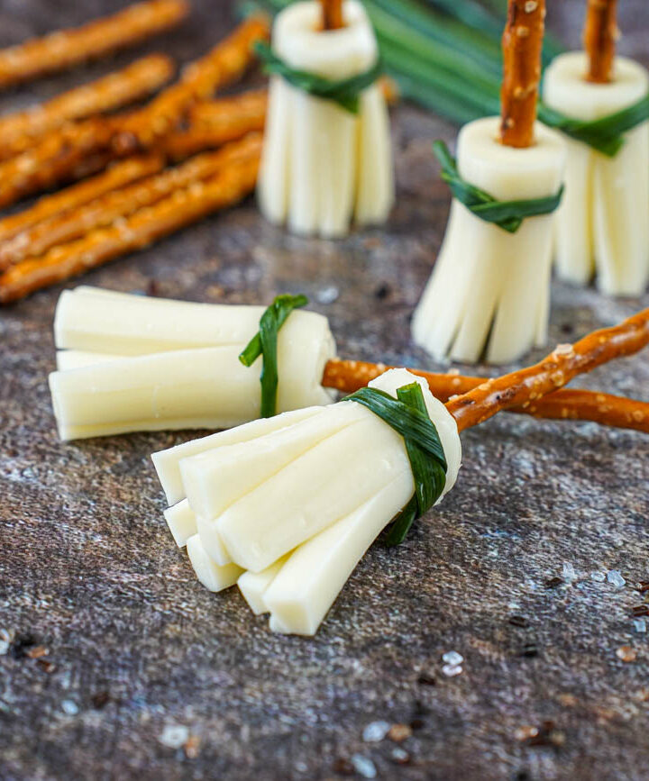 Two Pretzel Broomsticks on their side and three standing up in the background next to pretzel sticks and a bundle of chives.