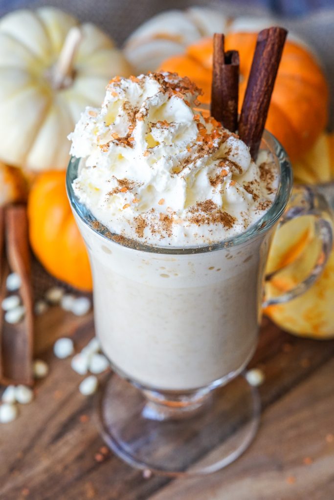 Pumpkin Spice White Hot Chocolate in a tall glass with stacked pumpkins in the background.