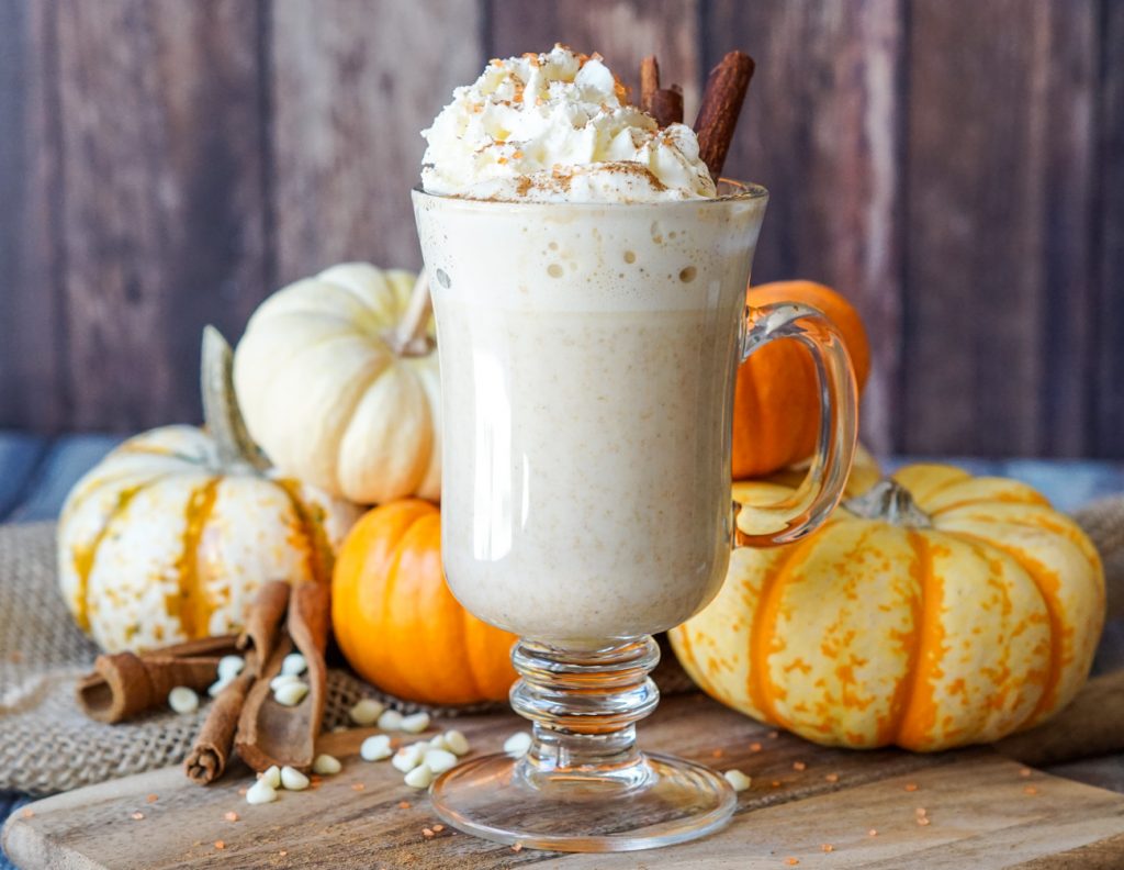 Pumpkin Spice White Hot Chocolate in a tall clear mug with mini pumpkins in the background.
