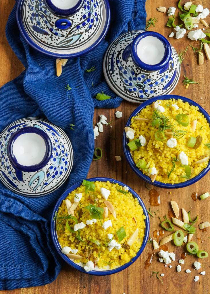 Aerial view of Yellow Couscous in two miniature clay blue/white tagines on a wooden platter with green onion, almond, and goat cheese.