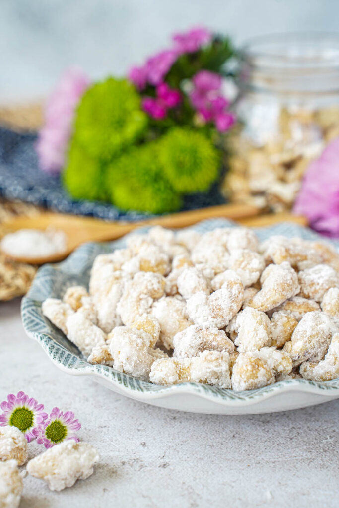 Side view of Sugar-Coated Cashew Nuts in a bowl with flowers and a jar of cashews in the background.