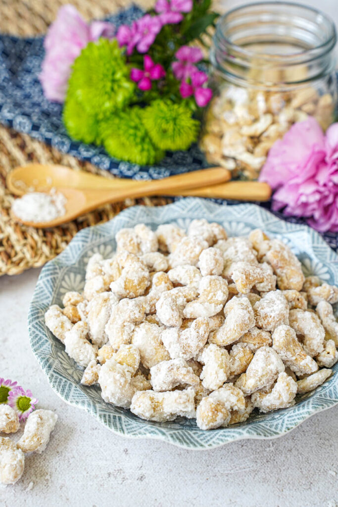 Sugar-Coated Cashew Nuts in a bowl with a jar of cashews, flowers, and two spoons in the background.