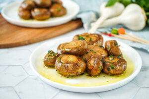 Champiñones al Ajillo (Spanish Garlic Mushrooms) on two white plates next to garlic and parsley.