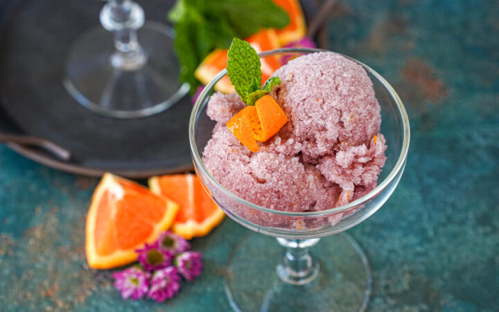Sorbete de Vino Tinto (Red Wine Sorbet) in a glass next to orange wedges and purple flowers.