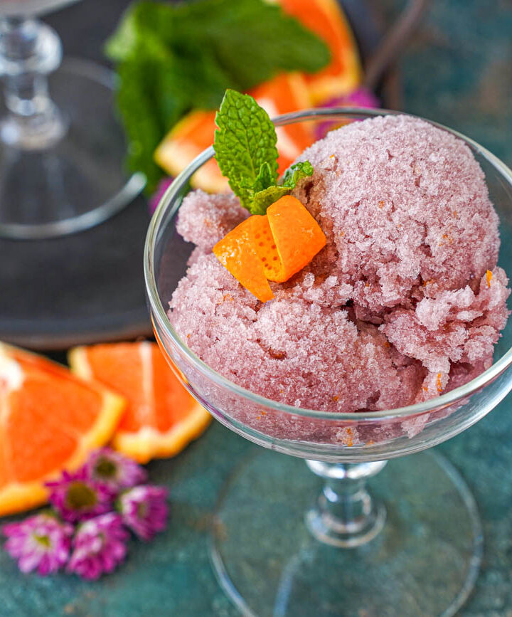 Sorbete de Vino Tinto (Red Wine Sorbet) in a glass next to orange wedges and purple flowers.
