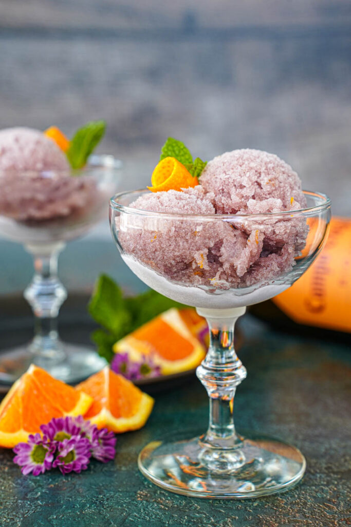 Side view of Sorbete de Vino Tinto (Red Wine Sorbet) in two glasses with orange wedges and purple flowers in the background.