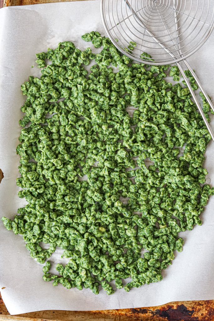 Aerial view of green Spätzli in a single layer on a parchment-lined baking sheet next to a steel strainer.
