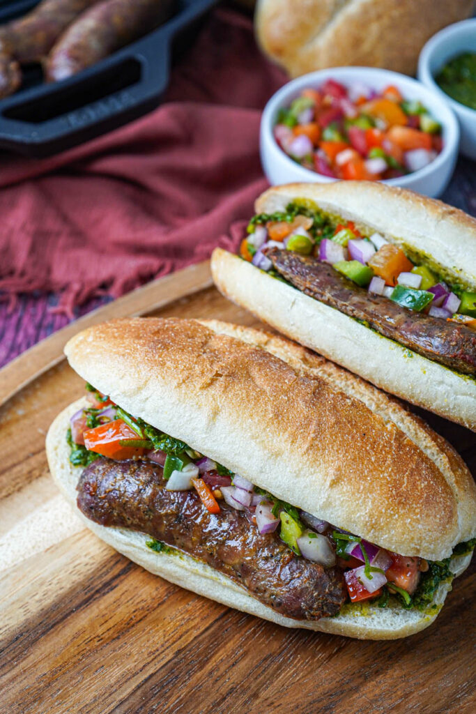 Two Choripán (Argentinian Chorizo Sandwiches) on a wooden board  with sausages, bread, and salsa criolla in the background.