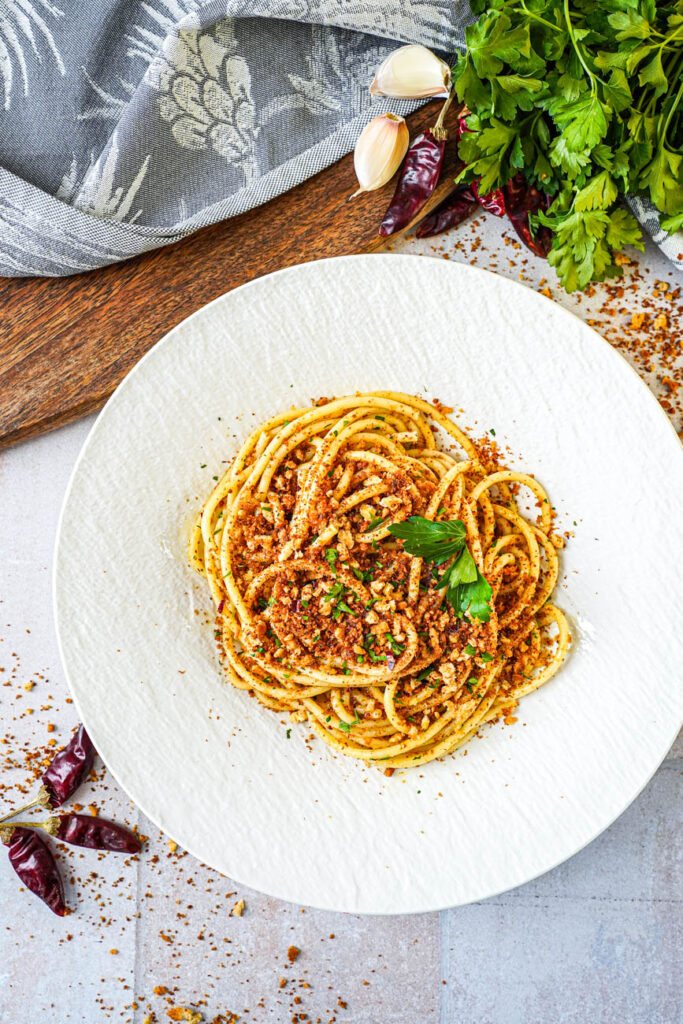 Aerial view of Pasta con la Mollica (Pasta with Breadcrumbs) in a white bowl next to dried chili peppers and fresh parsley.