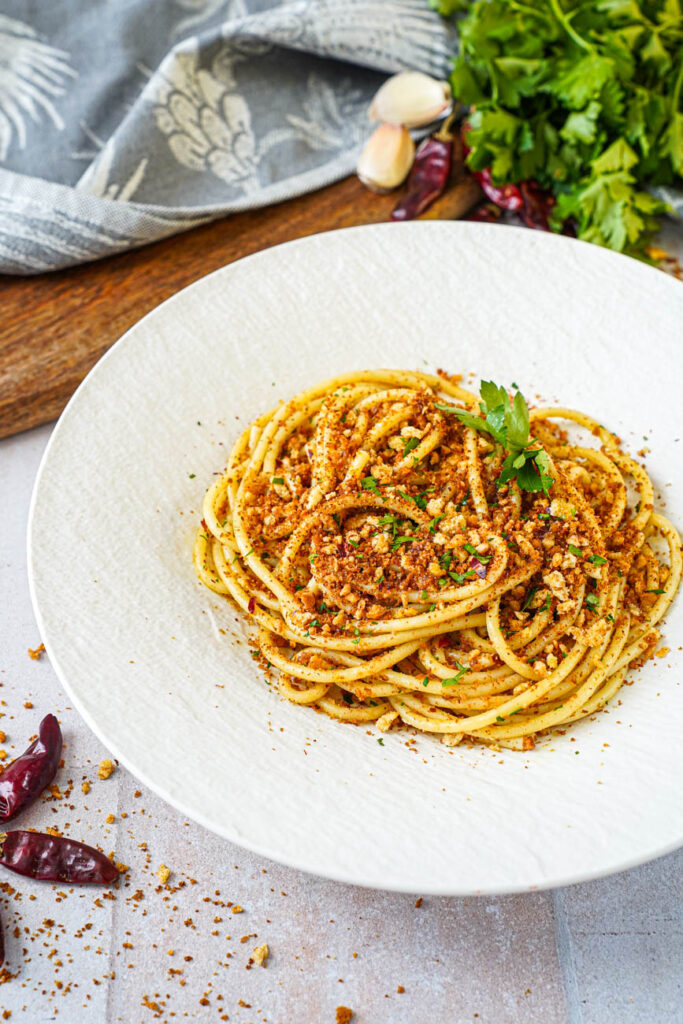 Close up of Pasta con la Mollica (Pasta with Breadcrumbs) with parsley, peppers, and garlic in the background.