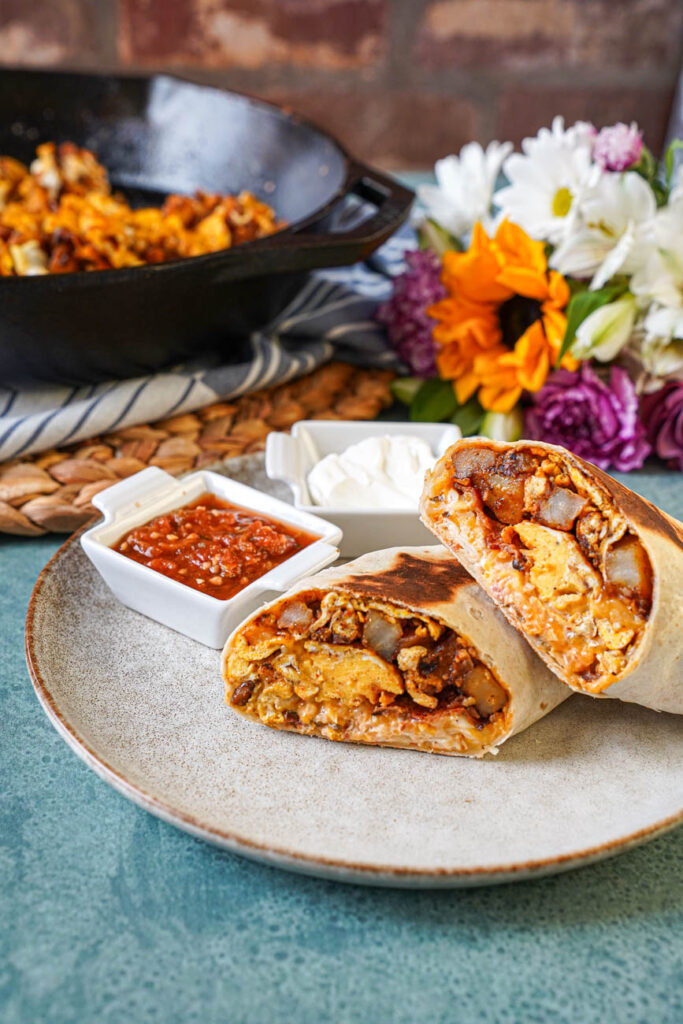Bacon Breakfast Burritos on a plate with flowers and a skillet of potatoes and eggs in the background.