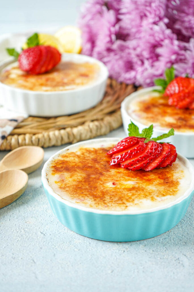 Side view of Strawberry Vanilla Yogurt Brûlée in blue and white ramekins with pink flowers in the background.