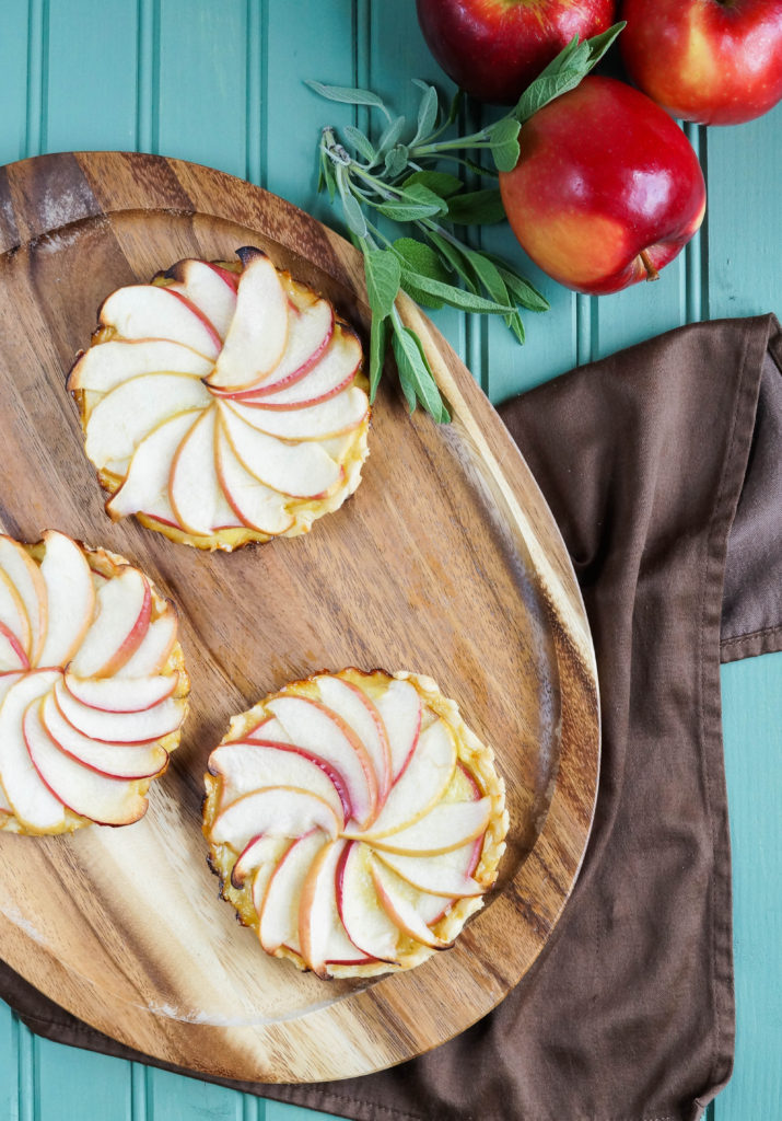 Aerial view of three Apple and Caramelized Onion Tarts on a wooden platter next to a brown towel, bunch of sage, and three apples.