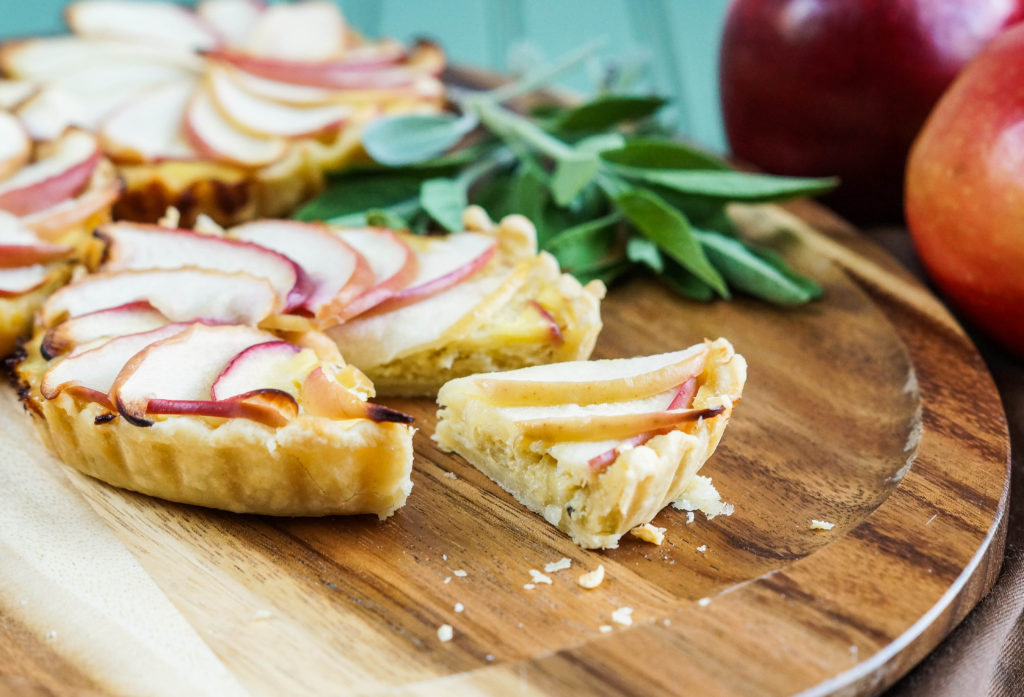 Three small Apple and Caramelized Onion Tarts on a wooden board with a small slice cut out. 