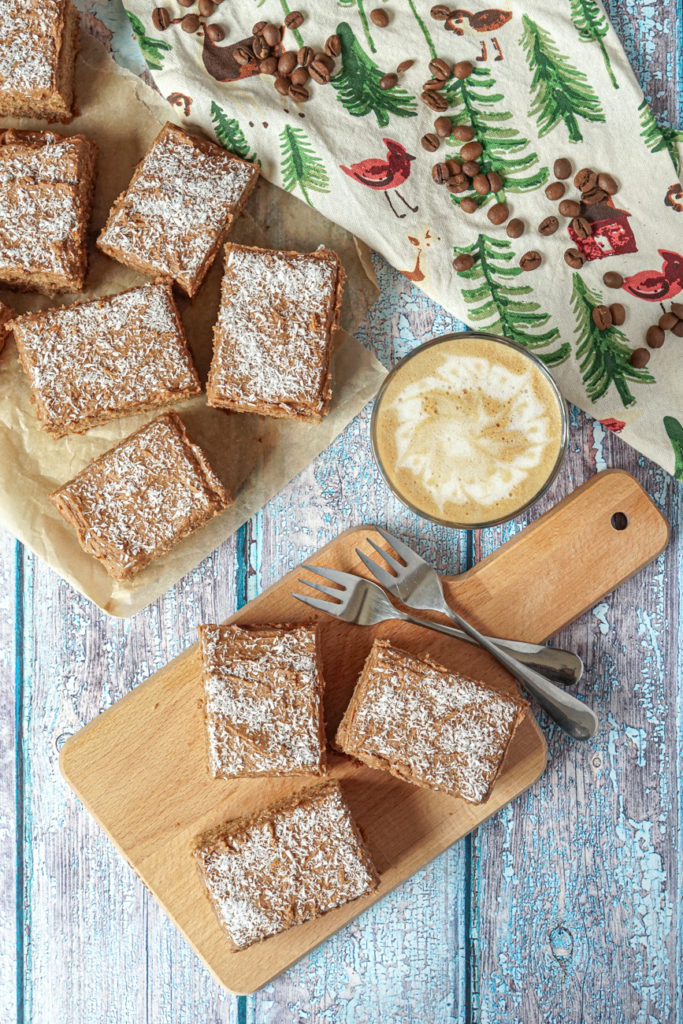 Aerial view of Kärleksmums (Swedish Coffee Cake)- three on a wooden board next to two silver forks and a latte. Six more slices are on a piece of brown parchment next to a towel with green trees and topped with a few coffee beans. 
