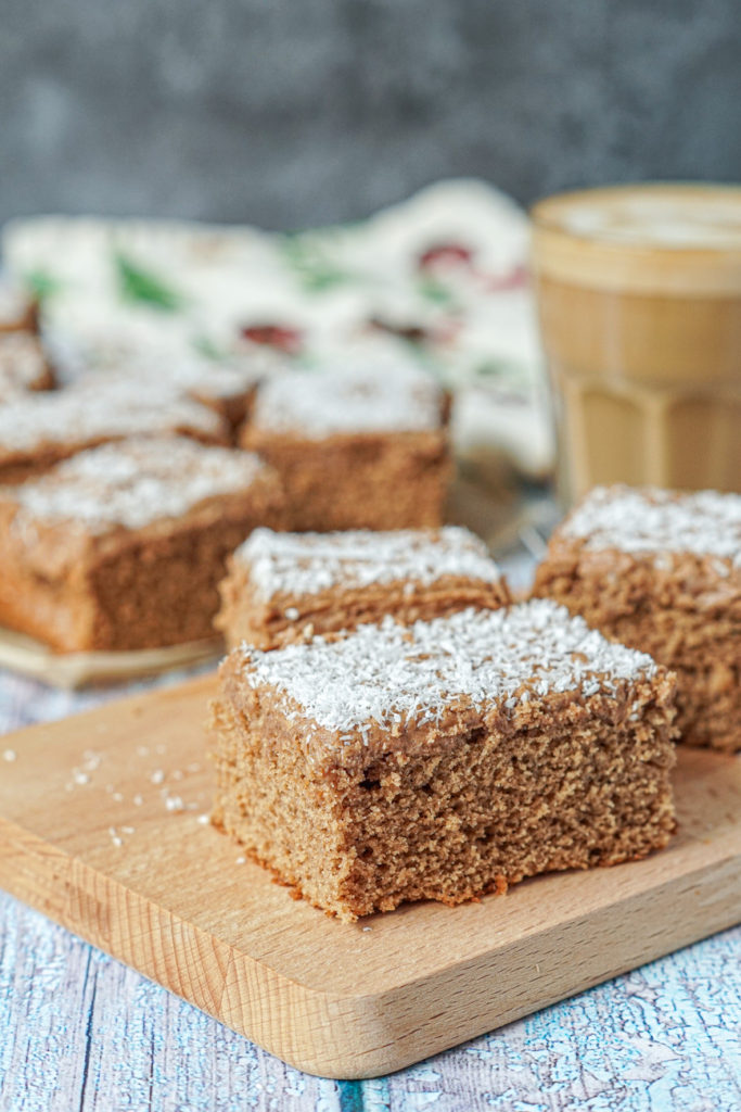 Side view of Kärleksmums (Swedish Coffee Cake) on a wooden board with a latte in a glass in the background.