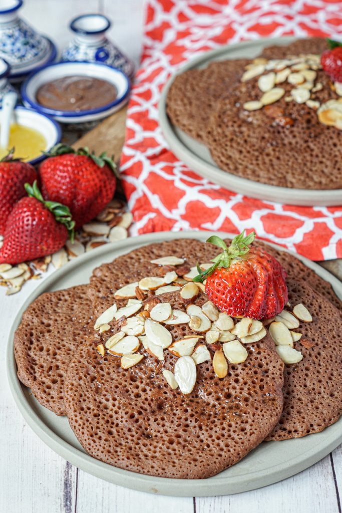 Side view of Chocolate Beghrir (Algerian Chocolate Honeycomb Pancakes) on two stone plates with strawberries and almonds.