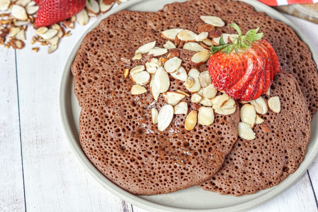 Chocolate Beghrir (Algerian Chocolate Honeycomb Pancakes) on a stone plate topped with toasted almonds and sliced strawberries.