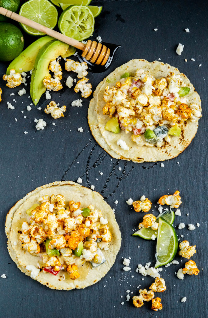 Aerial view of two Popcorn Tacos next to avocado slices, limes, and crumbled cheese.