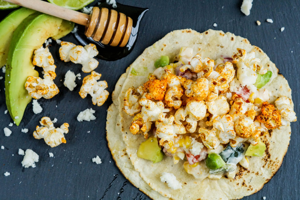 Close up of Popcorn Tacos next to a honey dipper with drizzled honey and two avocado slices.