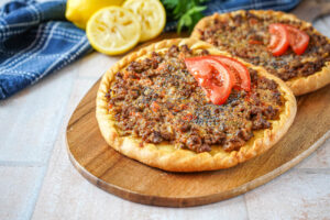 Two Aysh Abu Laham (Mini Saudi Pizzas) on a wooden board with lemon halves and parsley in the background.