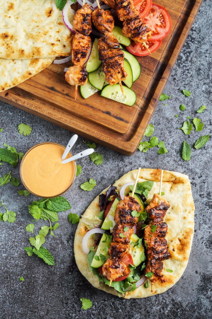 Aerial view of Grilled Chicken Naan Wraps with Roasted Red Pepper Tahini Sauce with more chicken, vegetables, and naan on a wooden cutting board.