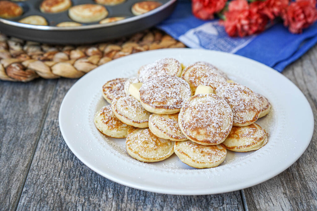 A pile of Poffertjes (Dutch Mini Pancakes) on a white plate with butter and powdered sugar.