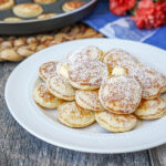 A pile of Poffertjes (Dutch Mini Pancakes) on a white plate with butter and powdered sugar.