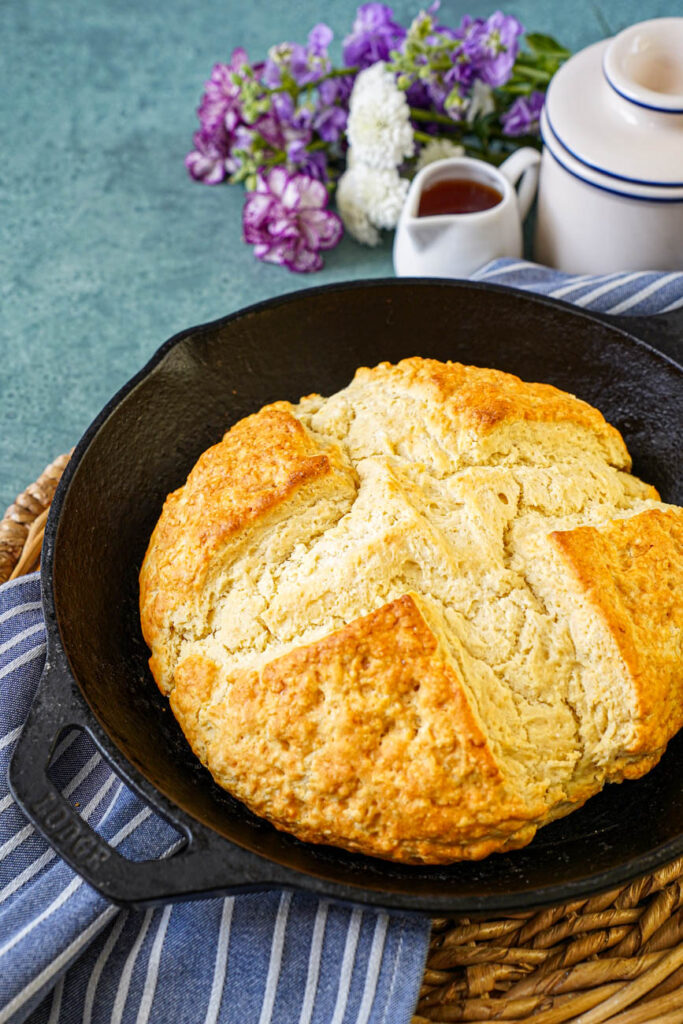 Close up of Australian Damper in a skillet next to a pitcher of golden syrup and purple and white flowers.