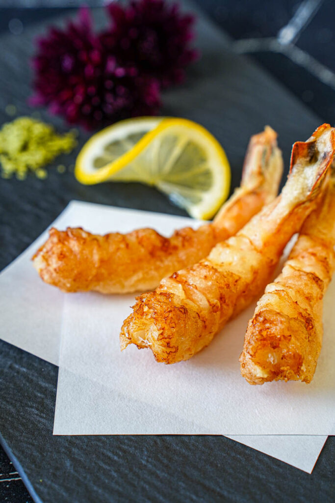 Close up of three Jumbo Shrimp Tempura on a black plate with a lemon slice, matcha salt, and dark purple flowers in the background.