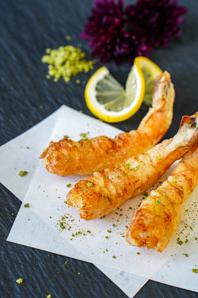 Close up of three Jumbo Shrimp Tempura on a black plate with a dusting of matcha salt.