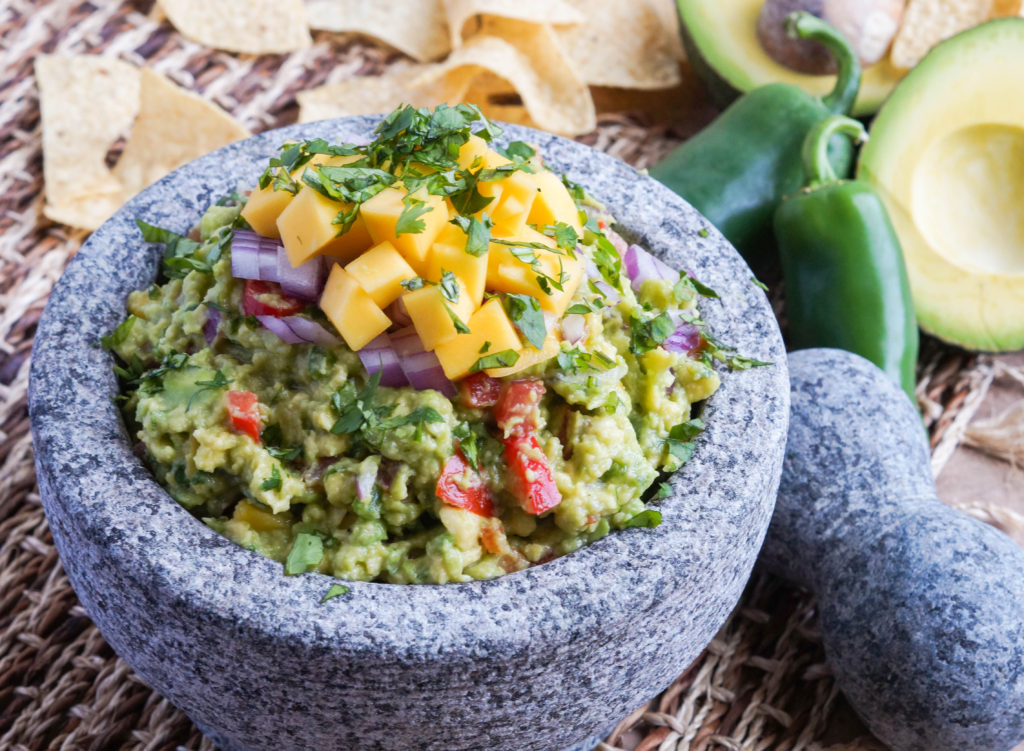 Mango Guacamole Mango Guacamole in a stone Molcajete next to jalapeños, avocados, and tortilla chips.
