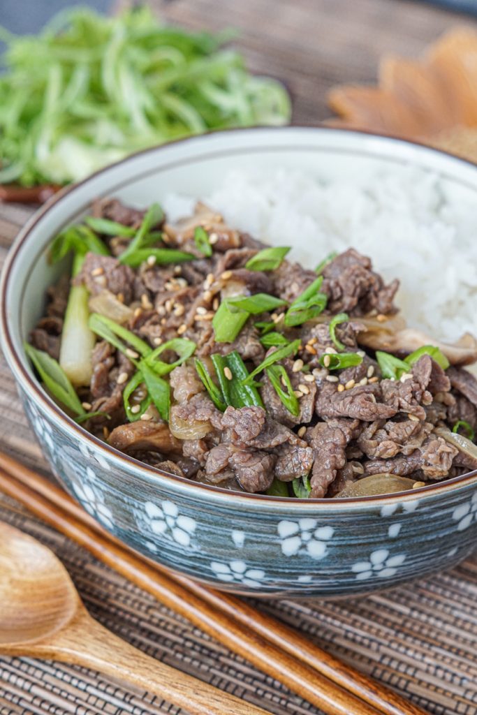 Close up of Bulgogi Dupbap (Korean Soy Garlic Beef over Rice) in a bowl next to wooden chopsticks and a spoon.