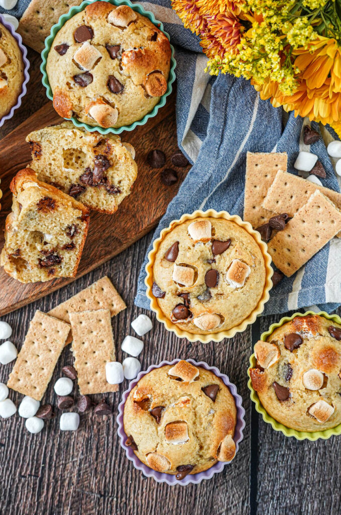 Aerial view of S'mores Muffins next to yellow flowers, graham crackers, mini marshmallows, and chocolate chips.